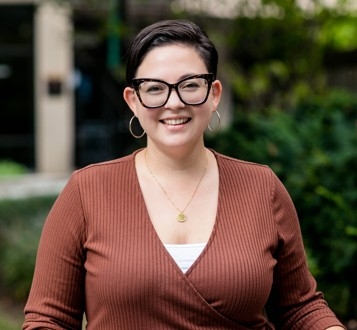 Marie McSweeney Anderson (she/they) wearing dark frame glasses and a brown sweater